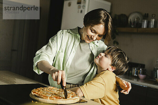 Junge umarmt lächelnde Mutter  die zu Hause in der Küche Pizza schneidet
