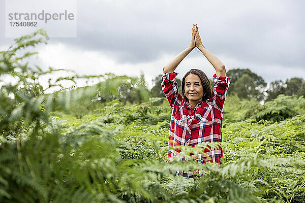 Smiling woman with clasped hands above head practicing yoga amidst plants at Cannock Chase