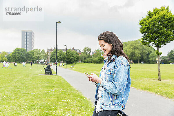 Lächelnde junge Frau in Jeansjacke  die im Park Textnachrichten auf dem Smartphone sendet