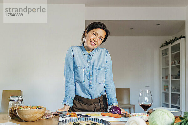Lächelnde Frau lehnt am Tisch mit Essen und Trinken in der heimischen Küche