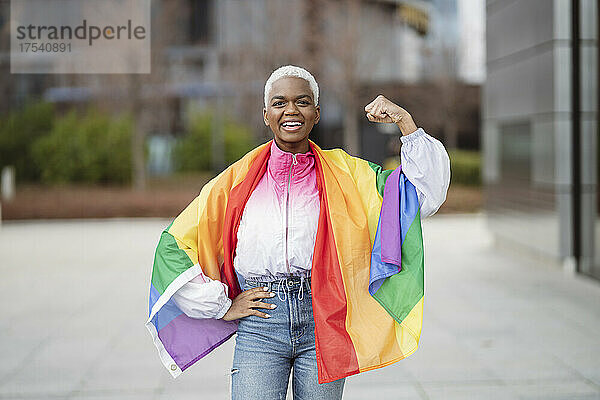 Young woman wrapped in rainbow flag flexing muscle with hand on hip