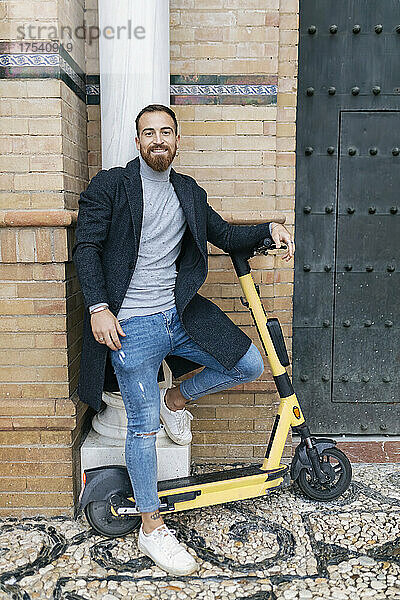 Happy bearded man with push scooter leaning on column