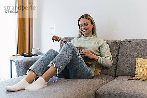 Smiling woman playing ukulele sitting on sofa at home
