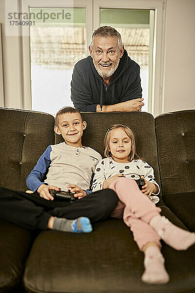 Mädchen und Junge spielen Videospiel mit Großvater  der sich zu Hause auf das Sofa lehnt