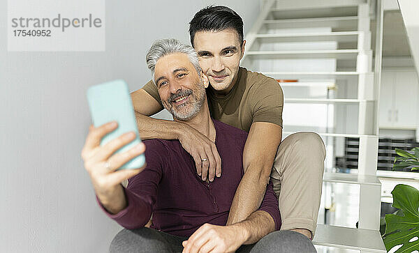 Smiling man taking selfie with husband through mobile phone at home