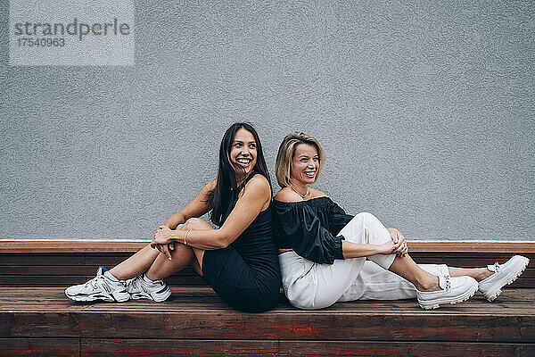 Lächelnde Freunde sitzen Rücken an Rücken auf einer Bank vor der Wand