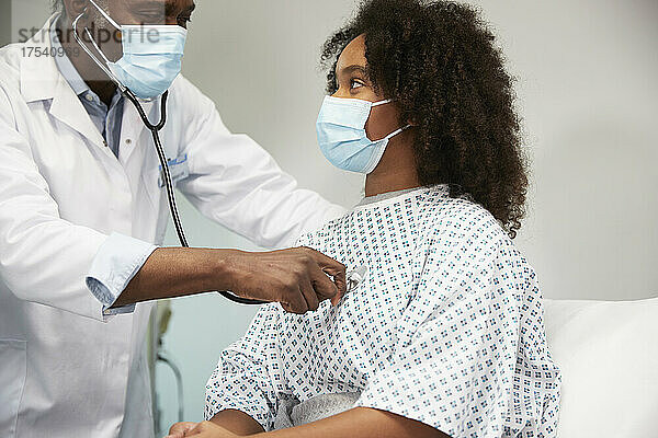 Male doctor with protective face mask examining patient with stethoscope at hospital