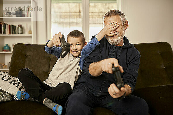 Enkel verdeckt die Augen seines Großvaters  der zu Hause ein Videospiel spielt