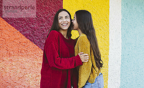 Young woman whispering in sister's ear by colorful wall