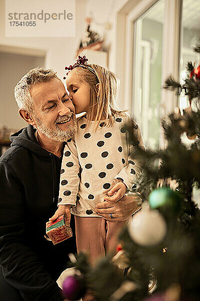 Cute girl holding gift kissing grandfather on christmas at home