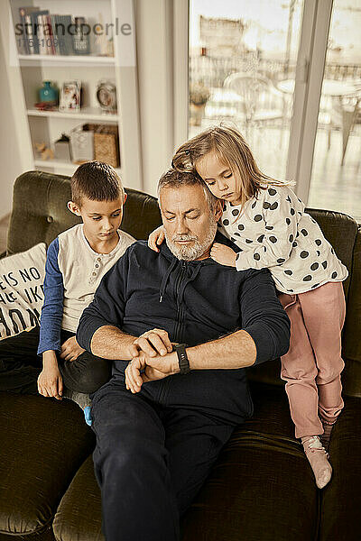 Junge und Mädchen sitzen mit Großvater und benutzen eine Smartwatch  die zu Hause auf dem Sofa sitzt