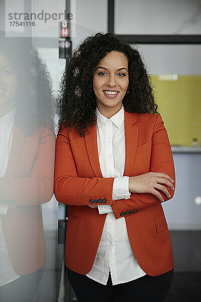Lächelnde Geschäftsfrau im orangefarbenen Anzug steht mit verschränkten Armen im Büro