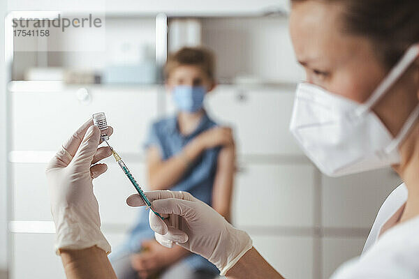 Krankenschwester bereitet den COVID-19-Impfstoff vor  mit einem Jungen im Hintergrund in der Mitte