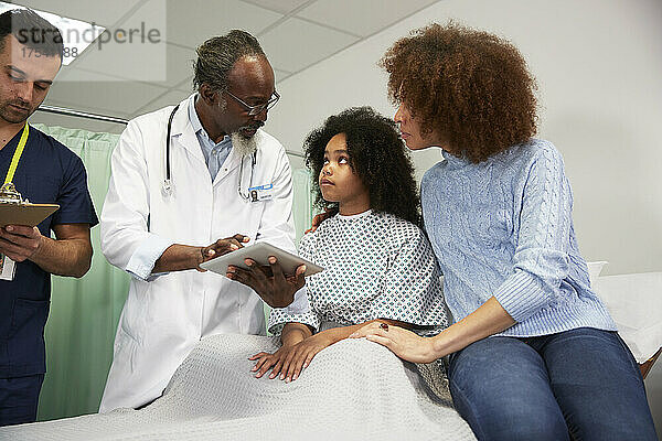 Patientin und Mutter hören dem Arzt zu  der im Krankenhaus einen Tablet-PC hält