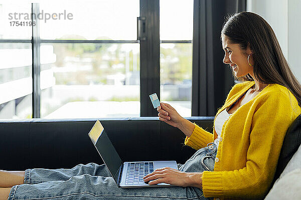 Glückliche Frau mit Kreditkarte beim Online-Shopping im Wohnzimmer