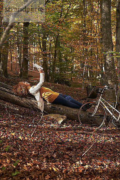 Woman holding book lying on log in autumn forest