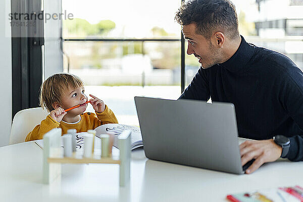 Vater spricht mit Sohn und arbeitet zu Hause am Laptop
