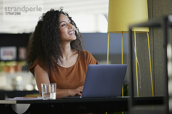 Glückliche Geschäftsfrau mit Laptop am Schreibtisch  die am Arbeitsplatz träumt