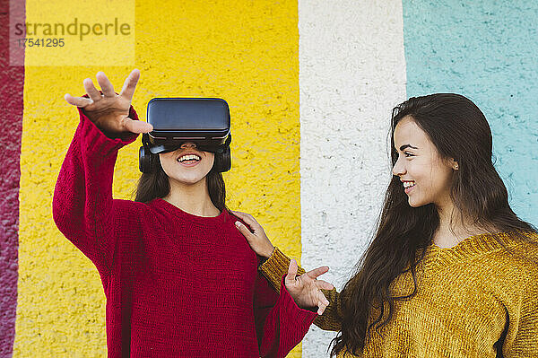 Woman smiling at sister wearing VR glasses and gesturing at colorful wall