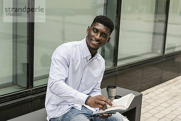 Lächelnder junger Mann mit Buch sitzt auf Bank