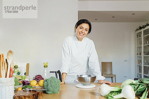 Lächelnder Koch lehnt zu Hause am Tisch mit grünem Gemüse