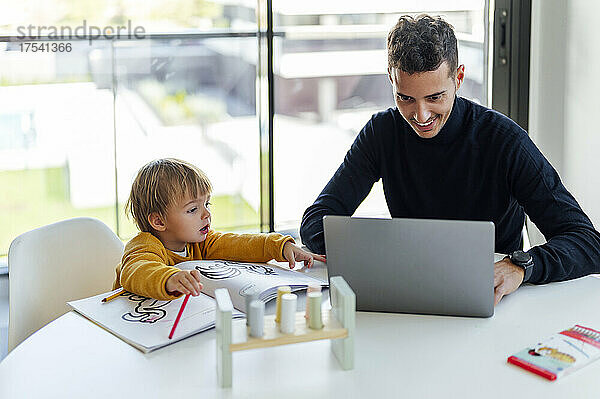 Lächelnder Geschäftsmann  der mit seinem Sohn zu Hause freiberuflich am Laptop arbeitet