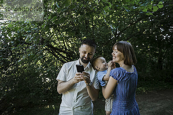 Smiling woman carrying son and looking at man showing smart phone