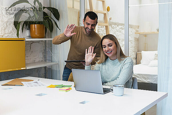 Lächelnde Kollegen grüßen per Videoanruf über Laptop am Schreibtisch im Studio