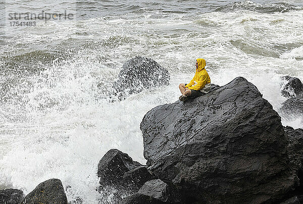 Young man wearing raincoat sitting on rock by waves splashing in sea  Rocha Da Relva  San Miguel Island  Azores  Portugal