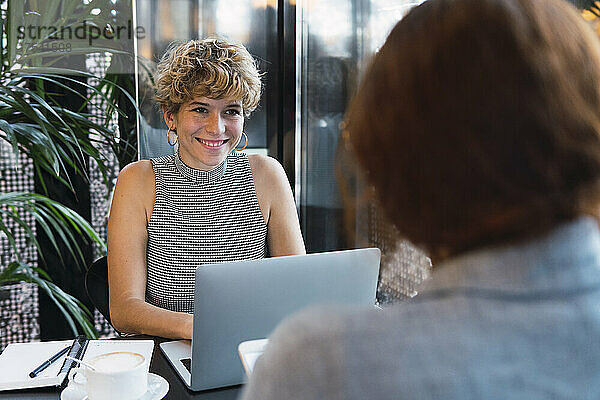 Young businesswoman with laptop smiling at colleague in coffee shop