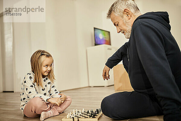 Girl looking at grandfather playing chess sitting at home
