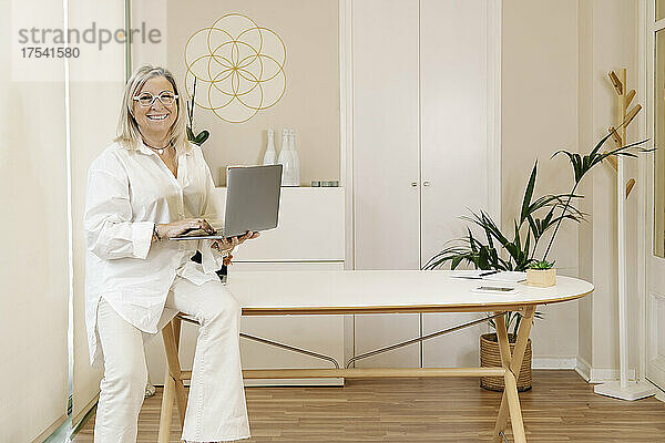 Smiling businesswoman holding laptop on desk at workplace