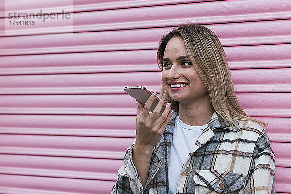 Young woman talking on speaker phone near pink shutter