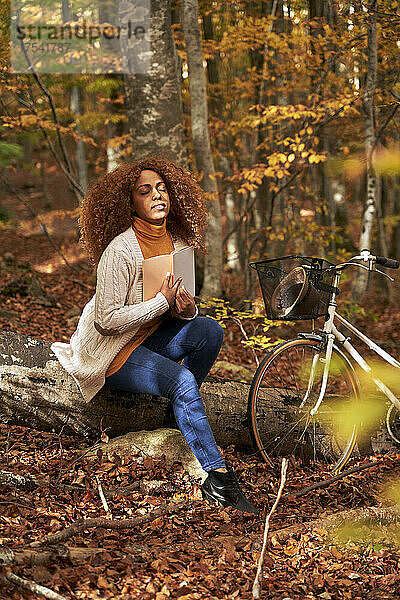 Frau mit geschlossenen Augen sitzt auf Baumstamm und hält Buch mit dem Fahrrad im herbstlichen Wald