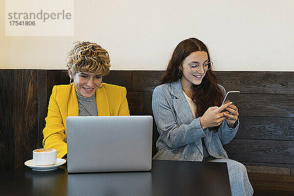 Lächelnde Geschäftsfrau  die von einem Kollegen am Laptop arbeitet und im Café ein Smartphone nutzt
