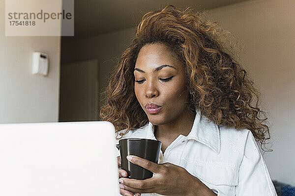 Junge Frau mit Laptop trinkt zu Hause Kaffee