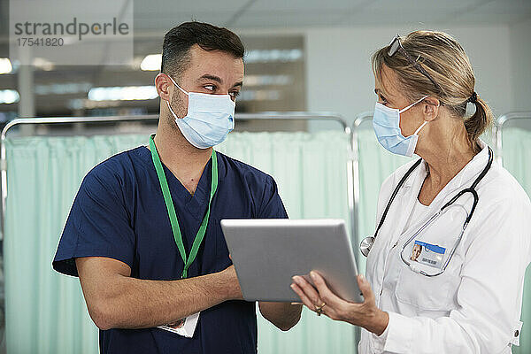 Krankenpfleger mit schützender Gesichtsmaske schaut den Arzt an  der im Krankenzimmer einen Tablet-PC hält