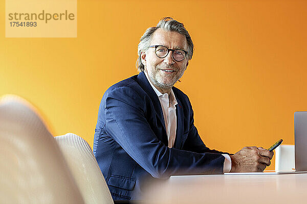 Smiling businessman with mobile phone at orange wall