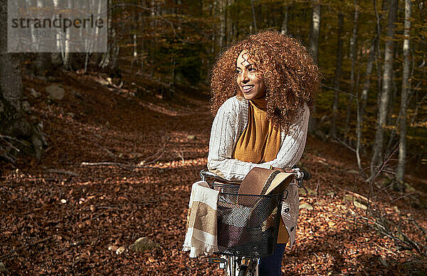 Lächelnde Frau stützt sich auf Fahrrad am Fußweg im Herbstwald