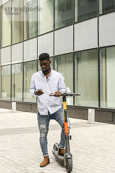 Junger Mann mit Tretroller und Mobiltelefon auf Fußweg