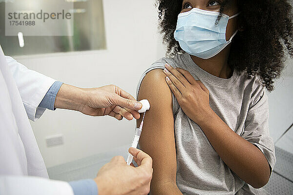 Doctor injecting COVID-19 vaccination to girl wearing protective face mask in medical room