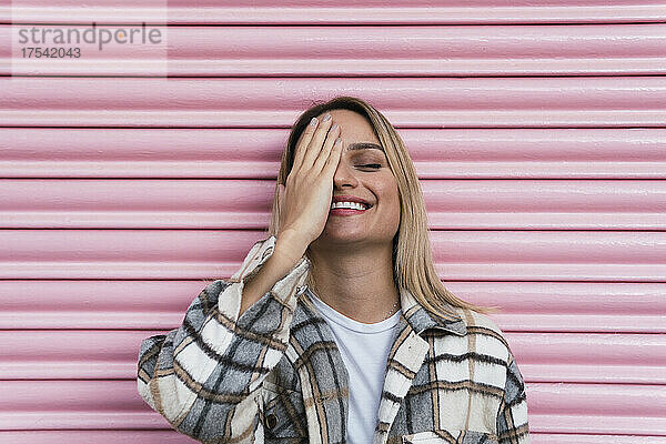 Smiling woman covering eye with hand in front of pink shutter