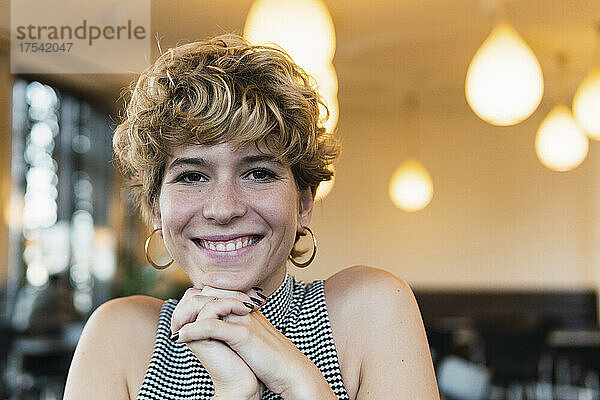 Glückliche junge Frau mit den Händen am Kinn lächelnd im Café