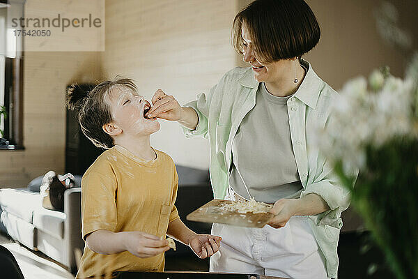 Smiling mother feeding grated cheese to son covered with flour on face at home