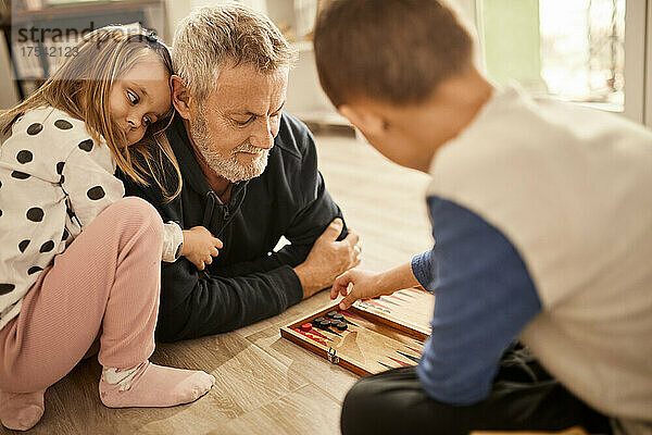 Enkelin umarmt Großvater und schaut Jungen zu  der zu Hause Backgammon spielt