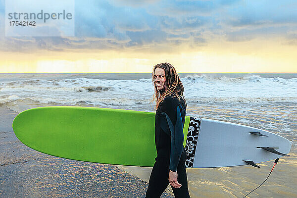 Lächelnder Surfer im Neoprenanzug  der mit Surfbrett am Meer spaziert