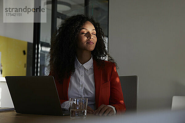 Geschäftsfrau sitzt mit geschlossenen Augen im Sonnenlicht am Arbeitsplatz