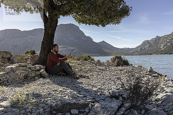 Mann sitzt auf einem Felsen neben einem Baum am Seeufer  Cuber-Staudamm  Mallorca  Balearen  Spanien