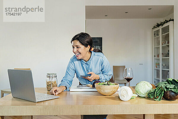 Lächelnde Frau hält ihr Smartphone bei einem Videoanruf mit einem Laptop zu Hause in der Hand