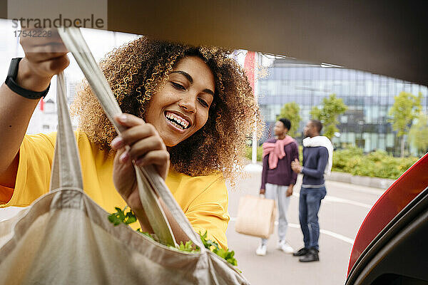 Lächelnde Frau lädt Einkaufstasche in den Kofferraum eines Autos und Freunde stehen im Hintergrund